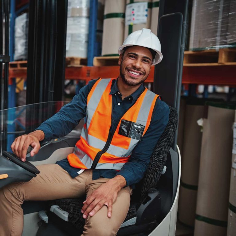 Portrait of happy young african forklift manager wearing safety vest and white hardhat transporting goods from one shelf to another while looking in camera in warehouse